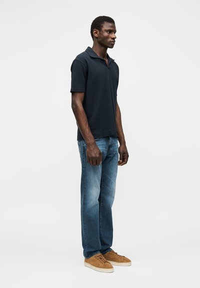 Homme debout en jean bleu, polo bleu marine foncé et baskets en daim marron sur un fond clair uni.