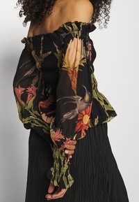 Blusa nera off-shoulder con corpino smockato e maniche, caratterizzata da motivi floreali colorati e colibrì.