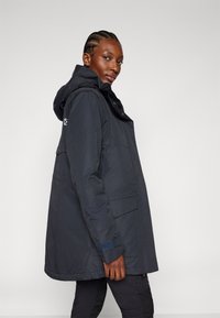 Marineblauwe waterdichte parka met een capuchon, voorzien van een rits aan de voorkant, grote zakken en gestructureerde manchetten. Gladde stof met minimale accenten.