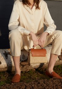 Personne en chemise et pantalon crème assise sur une bûche, tenant un sac à main tissé avec rabat en cuir marron et portant des mocassins marron.