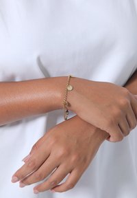 Bracciale in oro con piccoli ciondoli circolari, dalla finitura liscia e lucida, indossato su un polso su uno sfondo bianco.