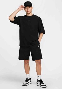Jauns vīrietis ar melnu Nike cepuri, pārāk lielu melnu Nike t-kreklu, melnām Nike šortiem, baltām Nike zeķēm un melnbaltiem Nike tenisa apaviem.