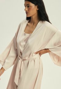 Jasnoróżowy satynowy szlafrok z szerokimi rękawami, związanym w talii oraz gładką teksturą, nałożony na sukienkę nocną w podobnym kolorze z subtelnymi wzorami.