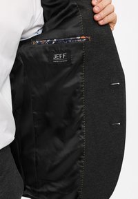 Mørk grå blazer med svart satengfòring, med en blå mønstret innerlomme, gul søm og en synlig merkevareetikett.