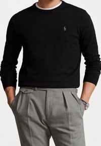 Jersey negro con cuello redondo y mangas largas, que presenta un pequeño logotipo bordado. Combinado con pantalones de vestir grises con pliegues.