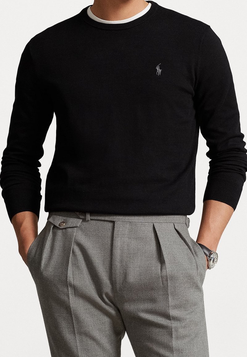 Jersey negro con cuello redondo y mangas largas, que presenta un pequeño logotipo bordado. Combinado con pantalones de vestir grises con pliegues.