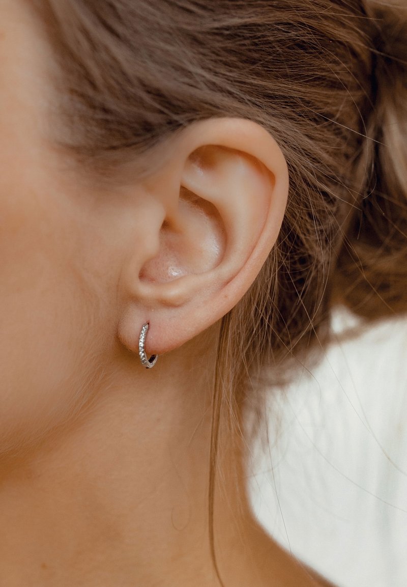 Gros plan sur l’oreille d’une femme aux cheveux châtain clair portant une petite boucle d’oreille en anneau argenté avec des pierres incrustées.