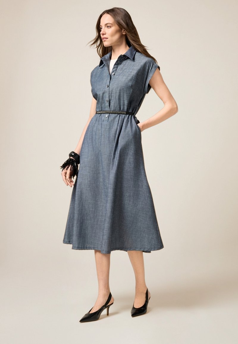Denim jurk met een kraaghals, kapmouwen en knoopsluiting aan de voorkant. Het heeft een tailleband en een vlammende rok. Zwarte puntige schoenen complementeren de outfit.