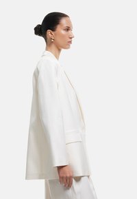 Kvinde med slicket hår i en knold, iført guldøreringe og en off-white oversized blazer, vendt mod højre foran en ensfarvet baggrund.