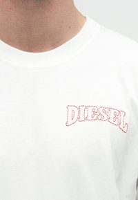Λευκό βαμβακερό t-shirt με κόκκινο κεντημένο λογότυπο "DIESEL" σε καμπυλωτό σχέδιο, με στρογγυλή λαιμόκοψη και απαλό, λείο ύφασμα.