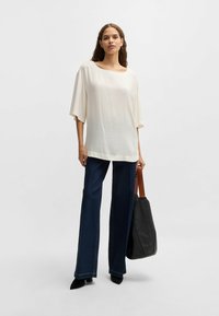 Top color crema a vestibilità ampia con maniche larghe, abbinato a jeans svasati blu scuro. La modella tiene una borsa tote nera con manici marroni.