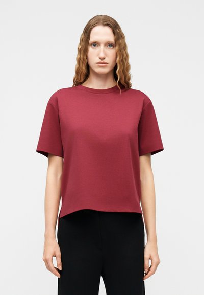 Femme aux cheveux ondulés portant un haut bordeaux lâche à manches courtes et un pantalon noir, debout devant un fond clair uni.