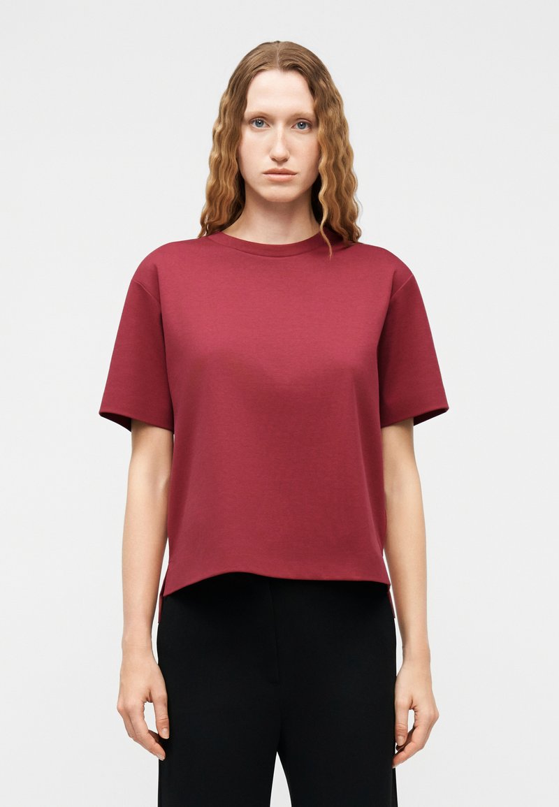 Femme aux cheveux ondulés portant un haut bordeaux lâche à manches courtes et un pantalon noir, debout devant un fond clair uni.