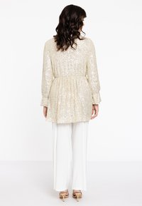 Goldfarbene Paillettentop mit plissierter Textur, taillierter Taille und langen Ärmeln, kombiniert mit weißen weit geschnittenen Hosen. Absatzsandalen runden den Look ab.
