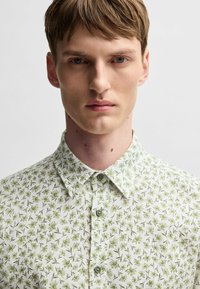 La camicia a maniche corte presenta una base bianca con un motivo di foglie verdi, un colletto a punta e bottoni verde chiaro. Tessuto testurizzato.