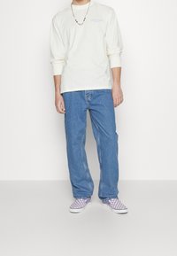 Persoon in een witte langemouwenshirt met klein logo, losse blauwe jeans, paars-wit geruite instapschoenen en een kralenketting.