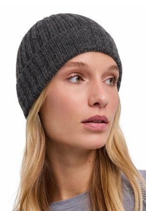 Junge Frau mit langen blonden Haaren, die eine dunkelgraue gerippte Strickmütze trägt und leicht nach rechts schaut, vor weißem Hintergrund.