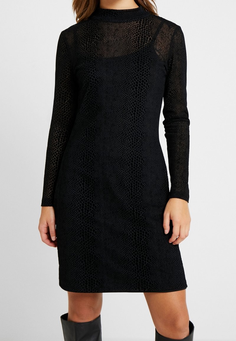 Svart långärmad klänning i genomskinligt, texturerat tyg med ett bikake-mönster, med hög halsringning och en figurnära silhuett.