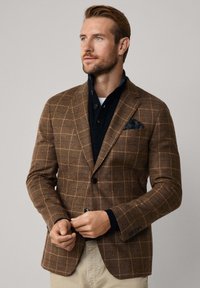 Braun karierter Blazer aus strukturiertem Stoff, mit Reverskragen, zwei Knöpfen und einem navyblauen Taschentuch mit Paisleymuster.