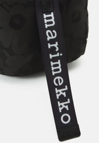 Borsa nera con un motivo floreale, dotata di una tracolla nera con la scritta "marimekko" in lettere bianche. Il materiale sembra testurizzato e resistente.
