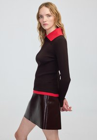 Młoda kobieta z blond włosami, ubrana w czarną bluzkę z długimi rękawami, z czerwoną kołnierzykiem i wykończeniem, w połączeniu z błyszczącą, ciemnobrązową skórzaną spódnicą.