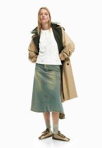 Mujer joven con un abrigo beige tipo trench, sudadera blanca con estampado floral, falda de mezclilla azul, calcetines grises y zapatillas beige, de pie con las manos en los bolsillos.