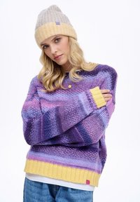 Pletený sveter s pásikmi v odtieňoch fialovej, ružovej a modrej, s žltým pruhovaným lemom. Doplnkom je svetlý pletený čiapka.