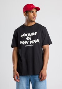Black cotton t-shirt with white graphic text reading "SOUNDS OF NEW YORK NY Social Club." Paired with a red cap, model faces sideways.