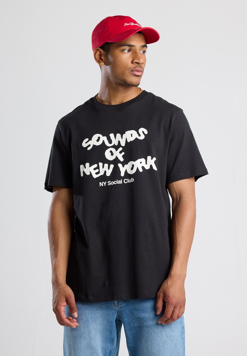 Black cotton t-shirt with white graphic text reading "SOUNDS OF NEW YORK NY Social Club." Paired with a red cap, model faces sideways.