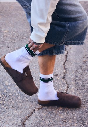 Személy farmer rövidnadrágot, fehér zöld csíkos zoknit, barna slip-on cipőt visel, és az egyik lábán lévő zoknit igazítja az aszfaltfelületen.