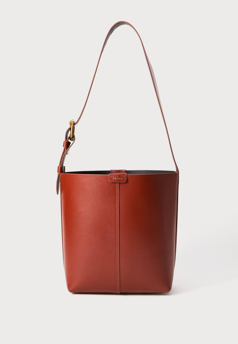 Hnědá kožená kabelka ve tvaru kbelíku s nastavitelným ramenním popruhem a zlatým přezkovým zapínáním, jednoduchý vertikální šev na přední straně.