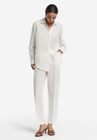 Hørhvid skjorte med knapper, løs pasform, lange ærmer og brystlomme, kombineret med matchende hørbukser med lige ben og brune hæle.