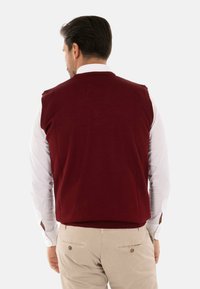 Maglione senza maniche bordeaux con una texture liscia, indossato sopra una camicia bianca a maniche lunghe. Il maglione presenta un collo rotondo e una vestibilità aderente.