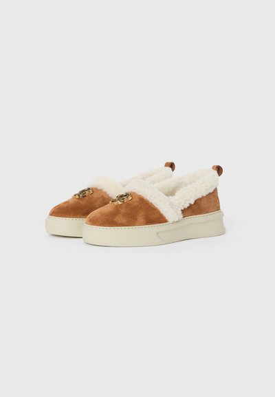 Brown suede slip-on shoes with a white fluffy lining, featuring gold hardware accents on the front and a light-colored rubber sole.