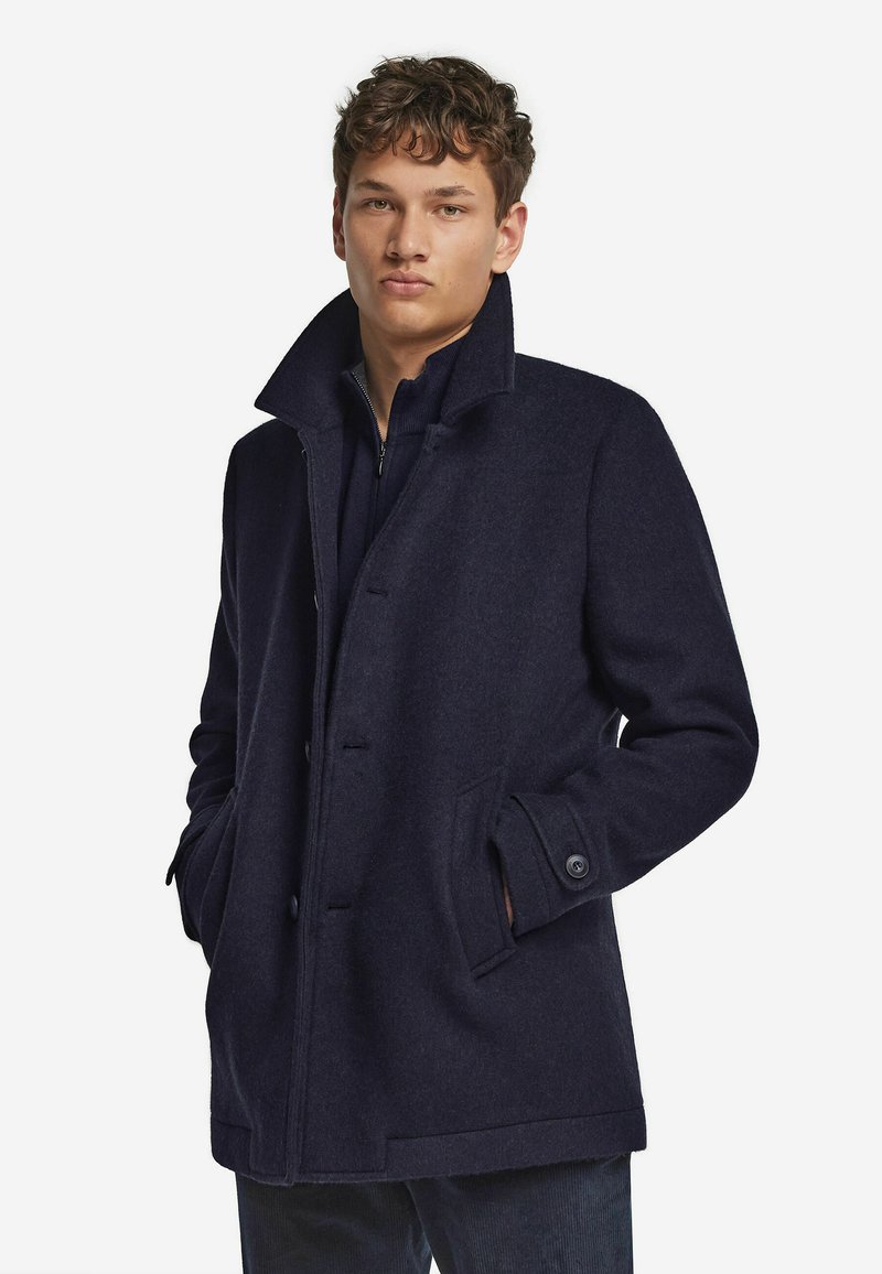 Marineblauer Wollmantel mit Knopfleiste, hohem Kragen und Taschen an den Seiten. Die glatte Textur und die taillierte Passform unterstreichen die Gesamt-Silhouette.