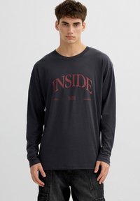 Joven con una camiseta negra de manga larga con texto y gráfico rojo que dice "INSIDE", combinada con pantalones cargo oscuros, posando frente a un fondo liso.