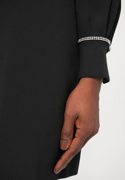 Manga de vestido negro con una textura suave, que presenta un detalle de rhinestones plateados a lo largo del puño. Mano posicionada cerca del vestido.
