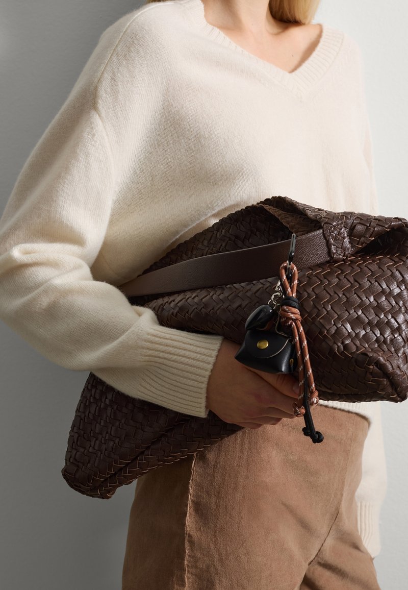 Persona con un suéter crema y pantalones marrones sosteniendo un gran bolso de mano de cuero trenzado marrón oscuro con accesorios de llavero adjuntos.
