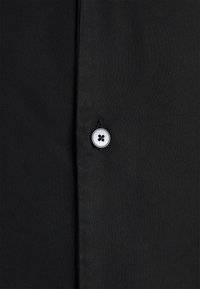 Tissu noir avec une surface lisse et texturée. Présente un bouton rond en argent avec un design à quatre trous, placé le long de la patte de boutonnage.