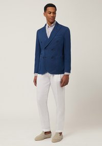 Blauer doppelt geknöpfter Blazer aus Baumwolle mit zwei Fronttaschen, kombiniert mit weißen Leinenhosen und hellgrauen Espadrilles. Gestreiftes Hemd darunter.