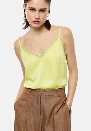 Giovane donna con capelli corti e scuri indossa una canotta giallo pallido con rifiniture in pizzo infilata in pantaloni marroni a vita alta, mani nelle tasche.