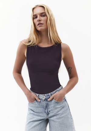 Blondynka z długimi włosami nosi czarny top bez rękawów i jasnoniebieskie jeansy, stoi z rękami w kieszeniach na białym tle.
