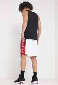 T-shirt sans manches noir, shorts blancs avec des rayures latérales rouges et noires, arborant un logo. Sneakers noires avec des accents rouges.