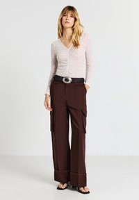 Top lungo a maniche lunghe in pizzo bianco con scollatura a V. Pantaloni cargo marroni a vita alta con grandi tasche, gambe larghe e risvolti. Cintura nera intrecciata con fibbia argentata.