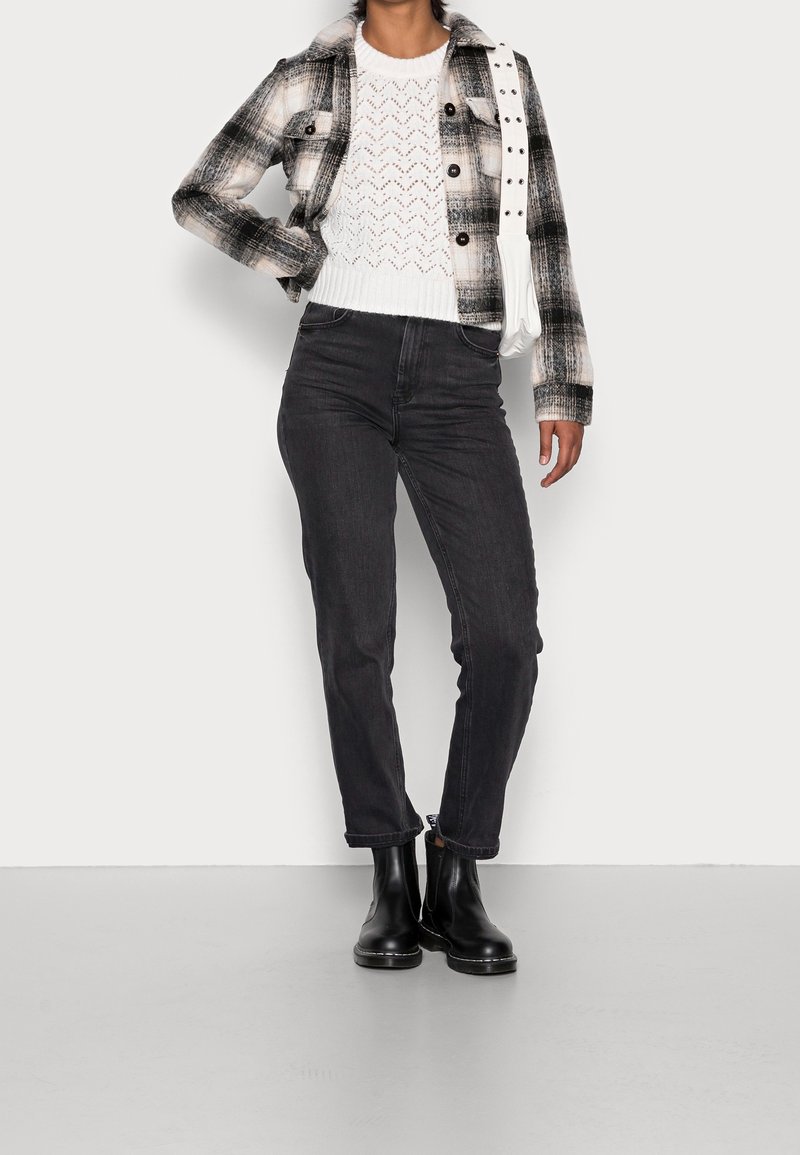 Schwarz-weiß kariertes Jackett mit Kragen, weißer gestrickter Pullover, schwarze Slim-Jeans und schwarze Stiefeletten. Trägt eine weiße Tasche.