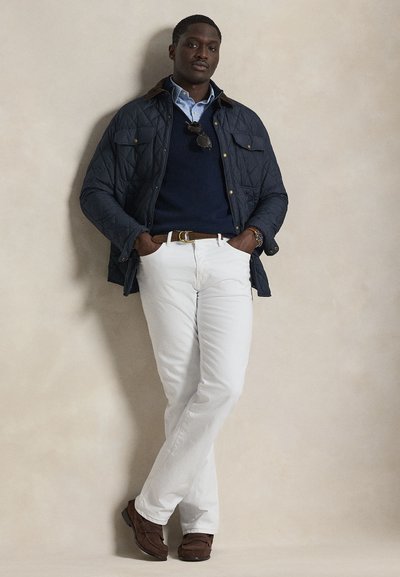 Μπλε μπλε μπουφάν με καφέ γιακά, μπλε πουλόβερ, ανοιχτό μπλε πουκάμισο, λευκό παντελόνι και καφέ παπούτσια. Χέρια στις τσέπες, στέκεται μπροστά σε μπεζ φόντο.