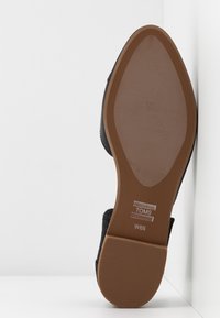 Semelle d'une chaussure TOMS noire taille W6.5 montrée sur un coin blanc, mettant en avant la semelle extérieure marron texturée et les motifs du talon.