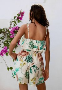 Jumpsuit met bloemenpatroon met een ruches bovenkant, verstelbare schouderbanden en een elastische taille. Gemaakt van lichtgewicht stof; hoofdzakelijk crèmekleurig met groene en roze accenten.