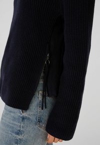 Marineblaue Strickjacke mit gerippter Textur, ausgestattet mit einem seitlichen Reißverschluss für eine verstellbare Passform und langen Ärmeln, kombiniert mit hellblauen Jeans.