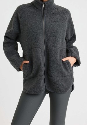 Person iført en mørkegrå fleecejakke med lynlås og hænderne i forlommerne, kombineret med grå ribstrikkede leggings mod en ensfarvet baggrund.